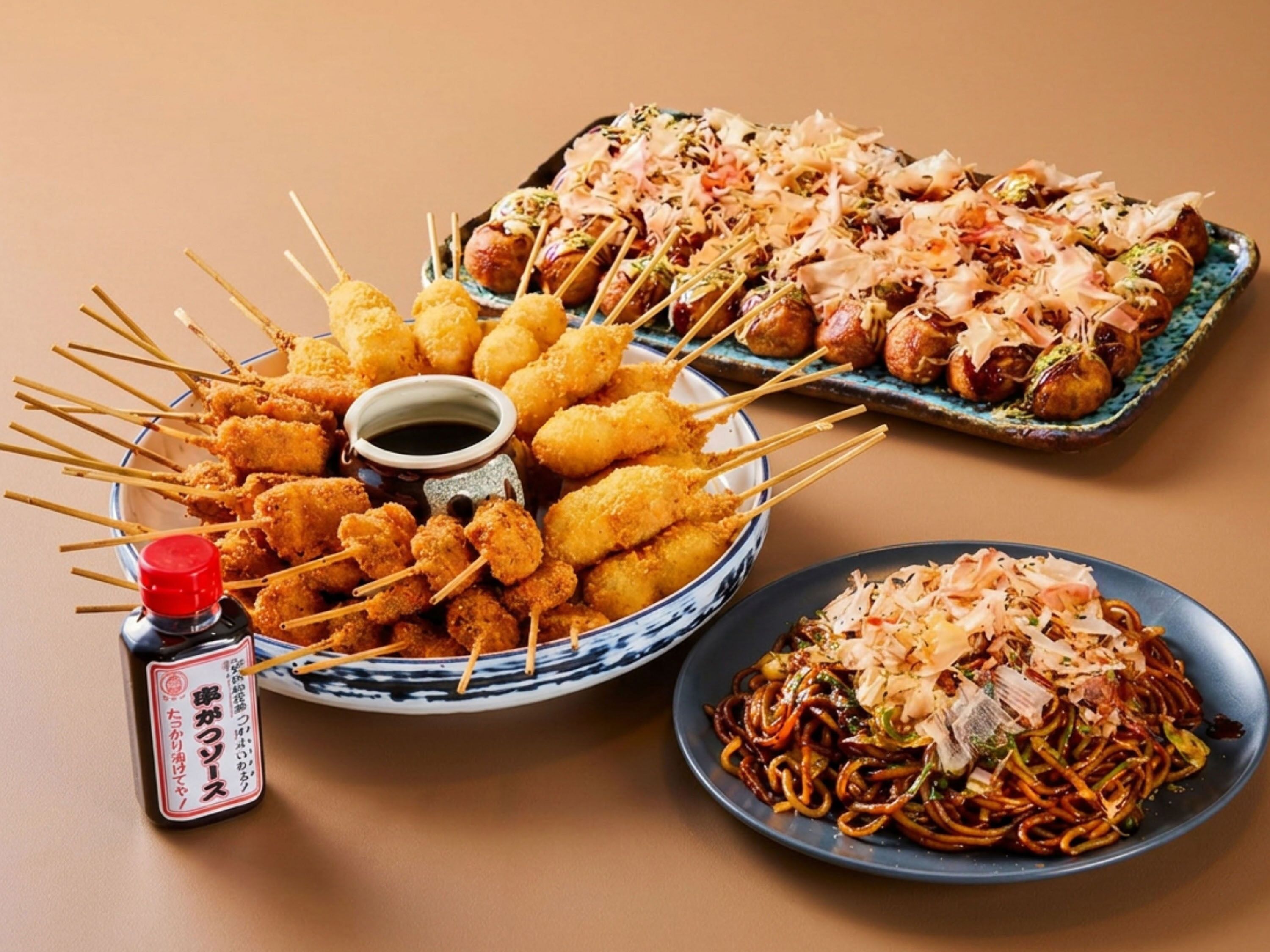 Ett urval av japanska rätter, inklusive spett, en skål med nudlar och ett fat med runda bitar garnerade med olika tillbehör.
