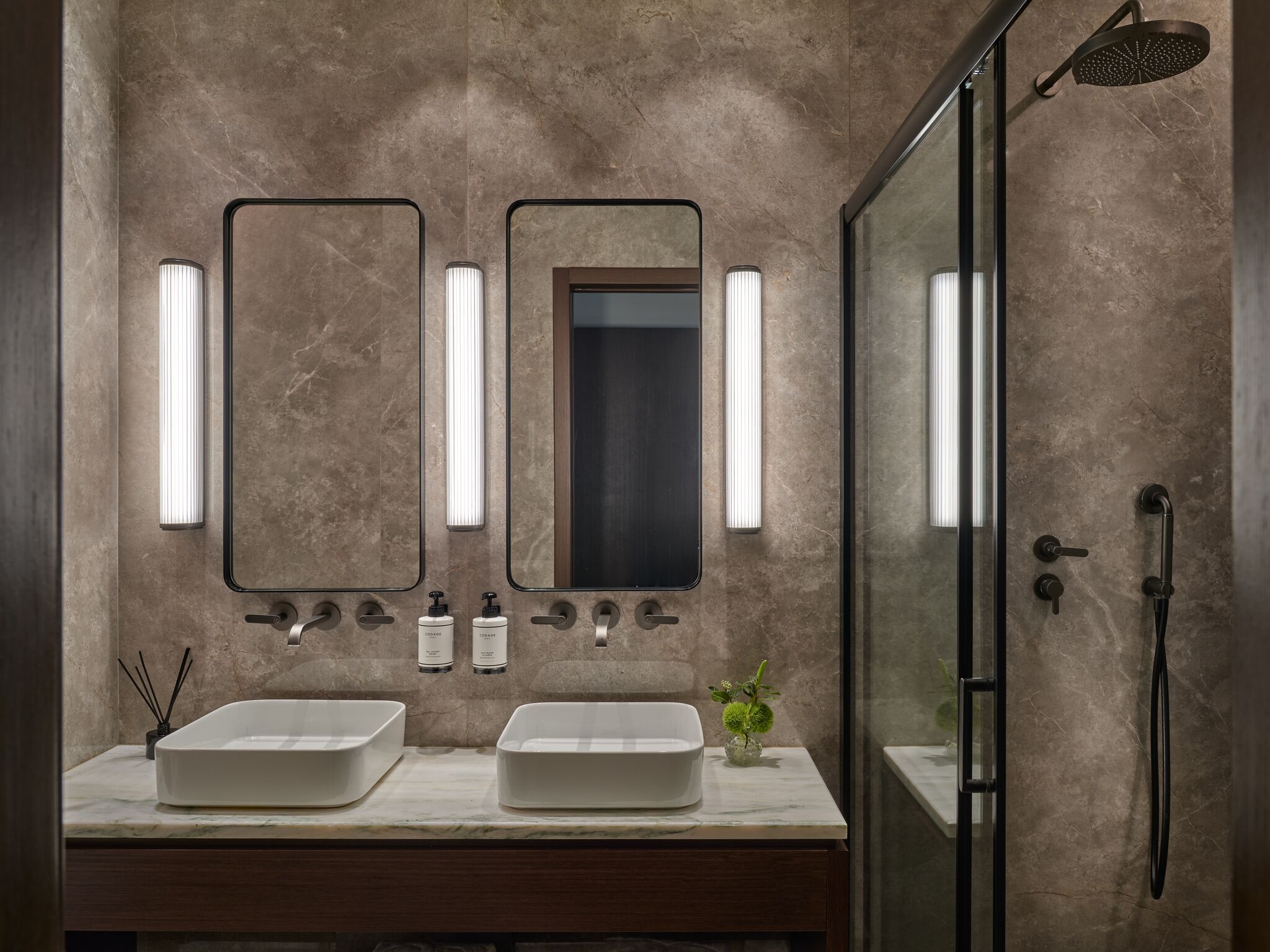 A modern bathroom with two sinks, a glass shower enclosure, and wall-mounted mirrors with vertical lights.
