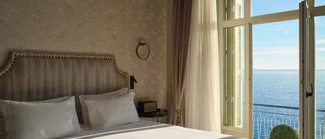 A large bed with a headboard, a bedside table, a clock, and a view of the sea through a balcony.