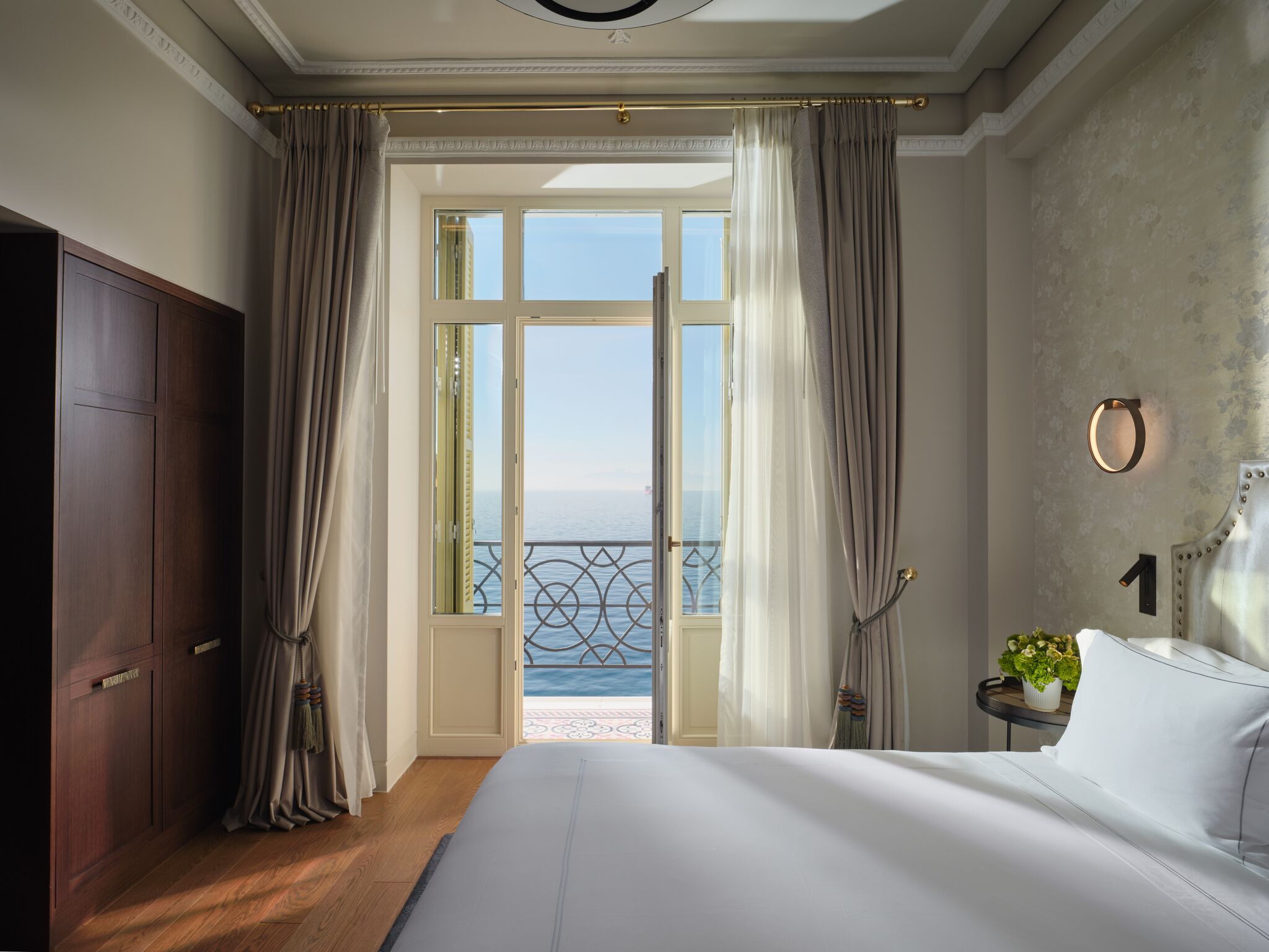 Un dormitorio con una cama grande, un armario, una mesita con una planta y vistas al mar a través de un ventanal.