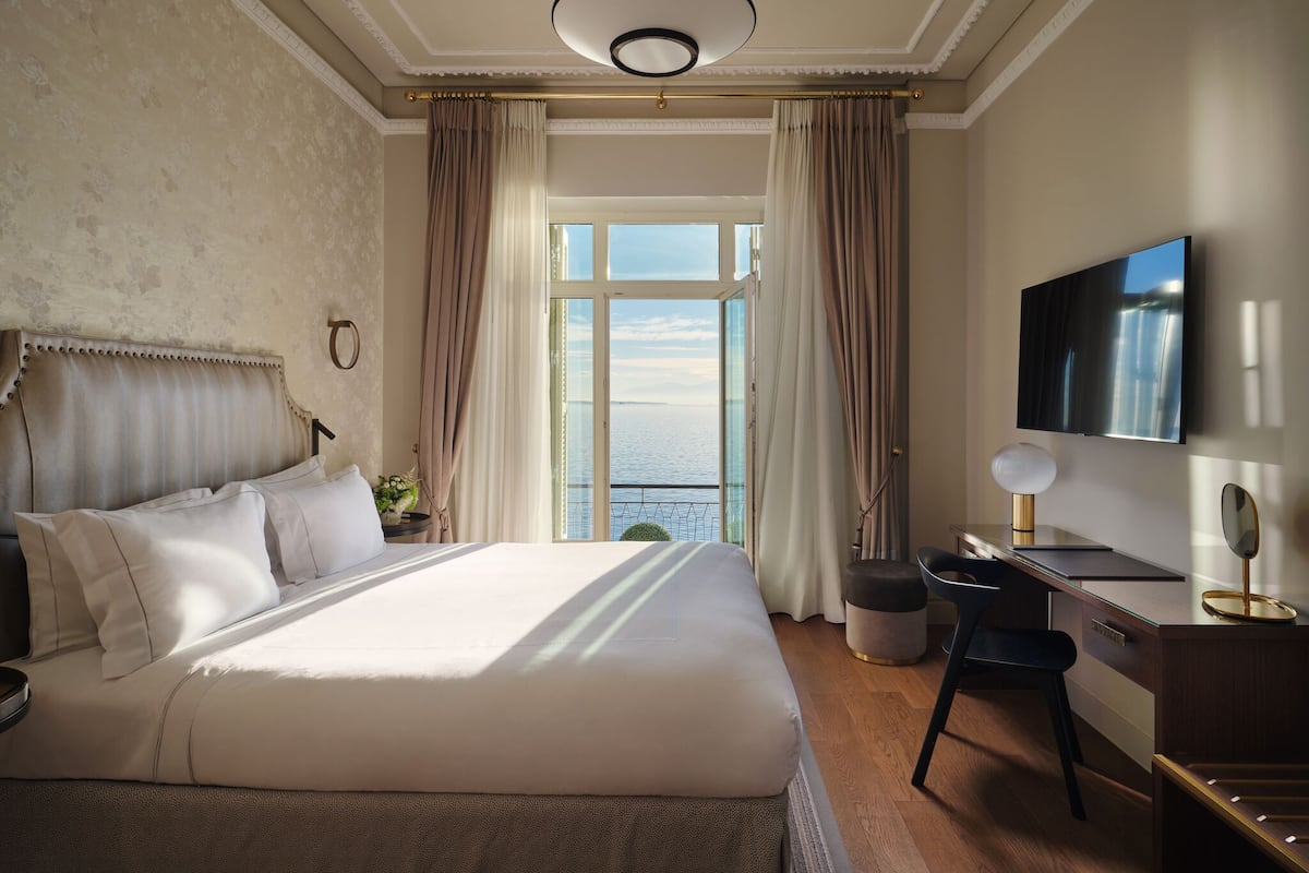 A hotel room with a large bed, a desk, a television, and a view of the sea.
