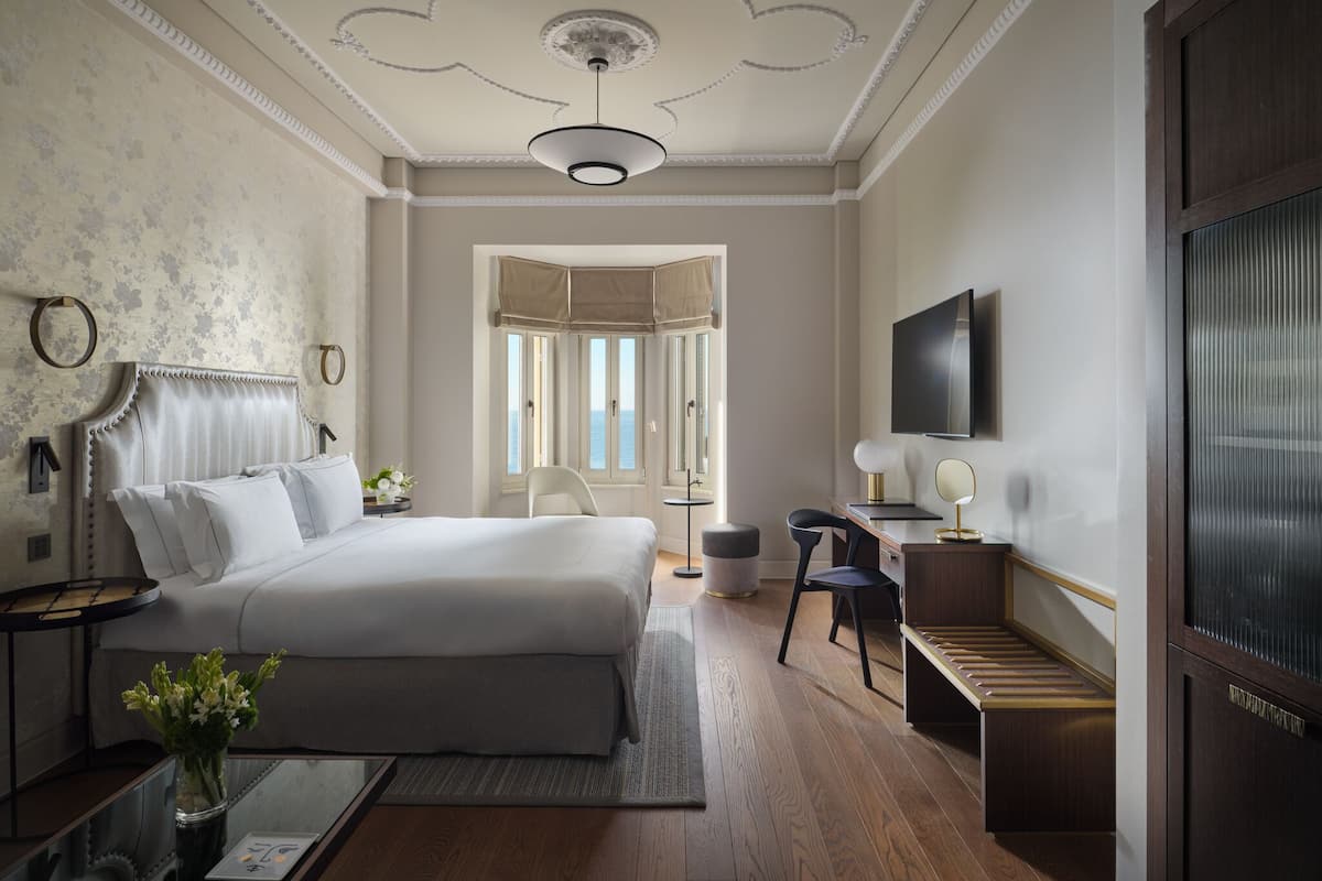 A hotel room with a bed, a desk, a chair, a TV, and a view of the sea through the windows.