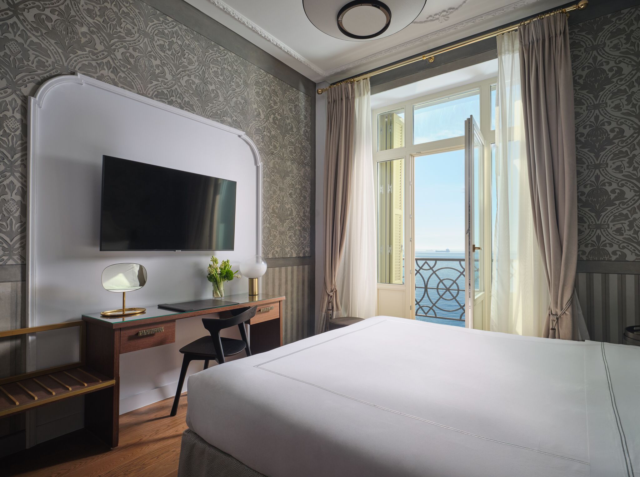 A hotel room with a bed, desk, TV, and a balcony with curtains.
