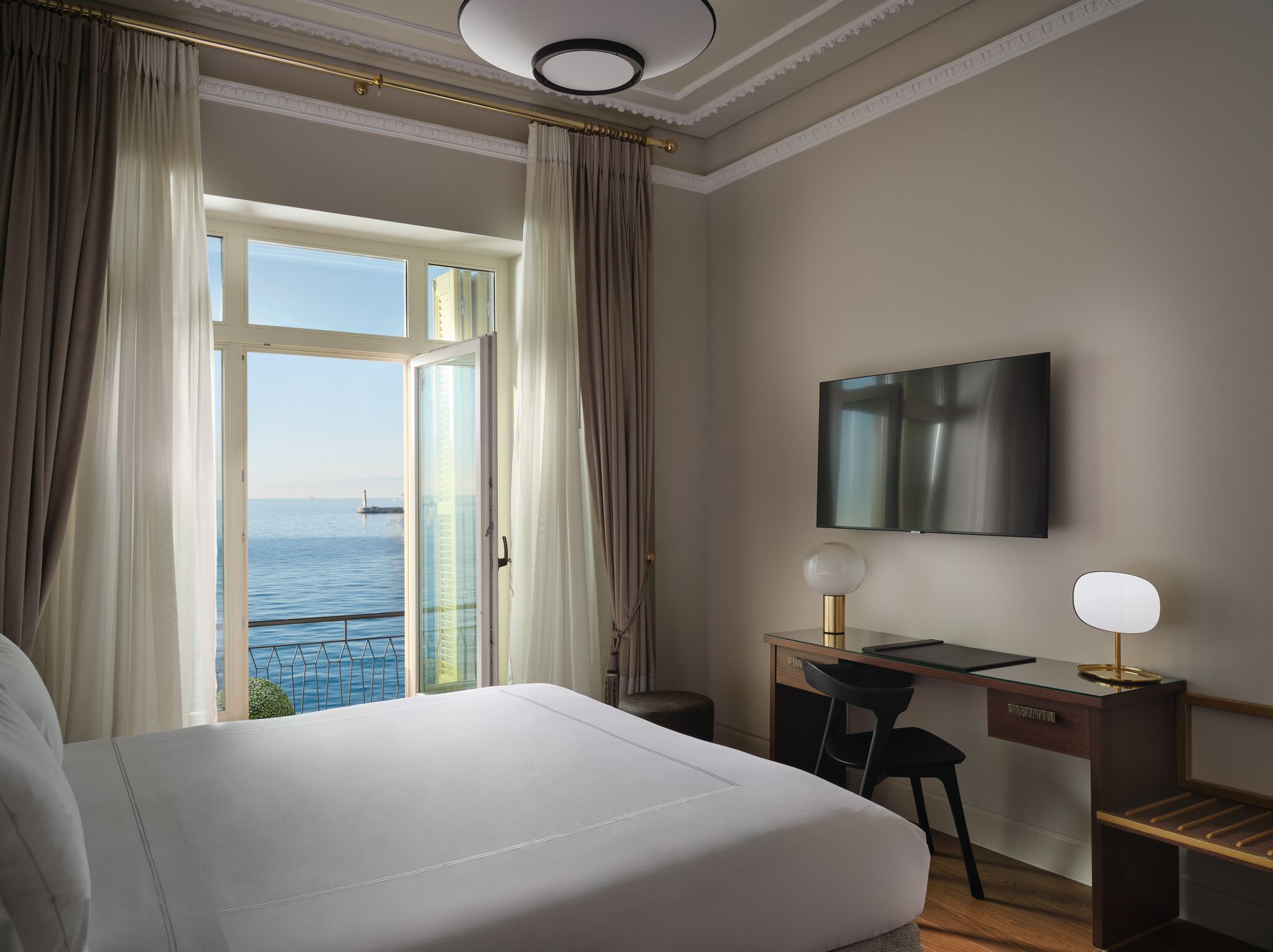 A hotel room with a large bed, a desk with a chair, a TV mounted on the wall, and a view of the sea through a large window.