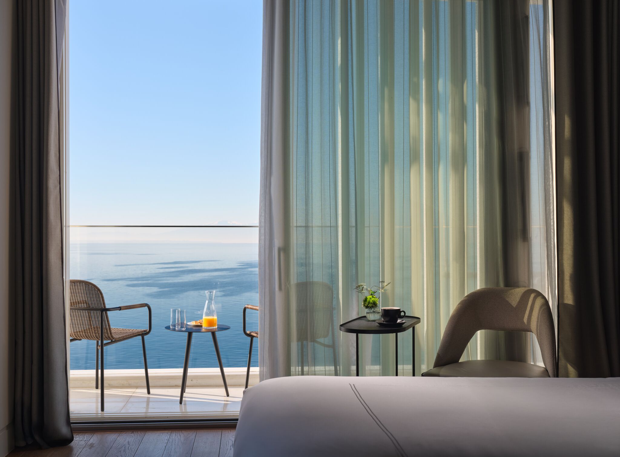 Een hotelkamer met balkon, een bed, twee stoelen, een klein tafeltje met een glas sinaasappelsap en een kop, en een bloevas.