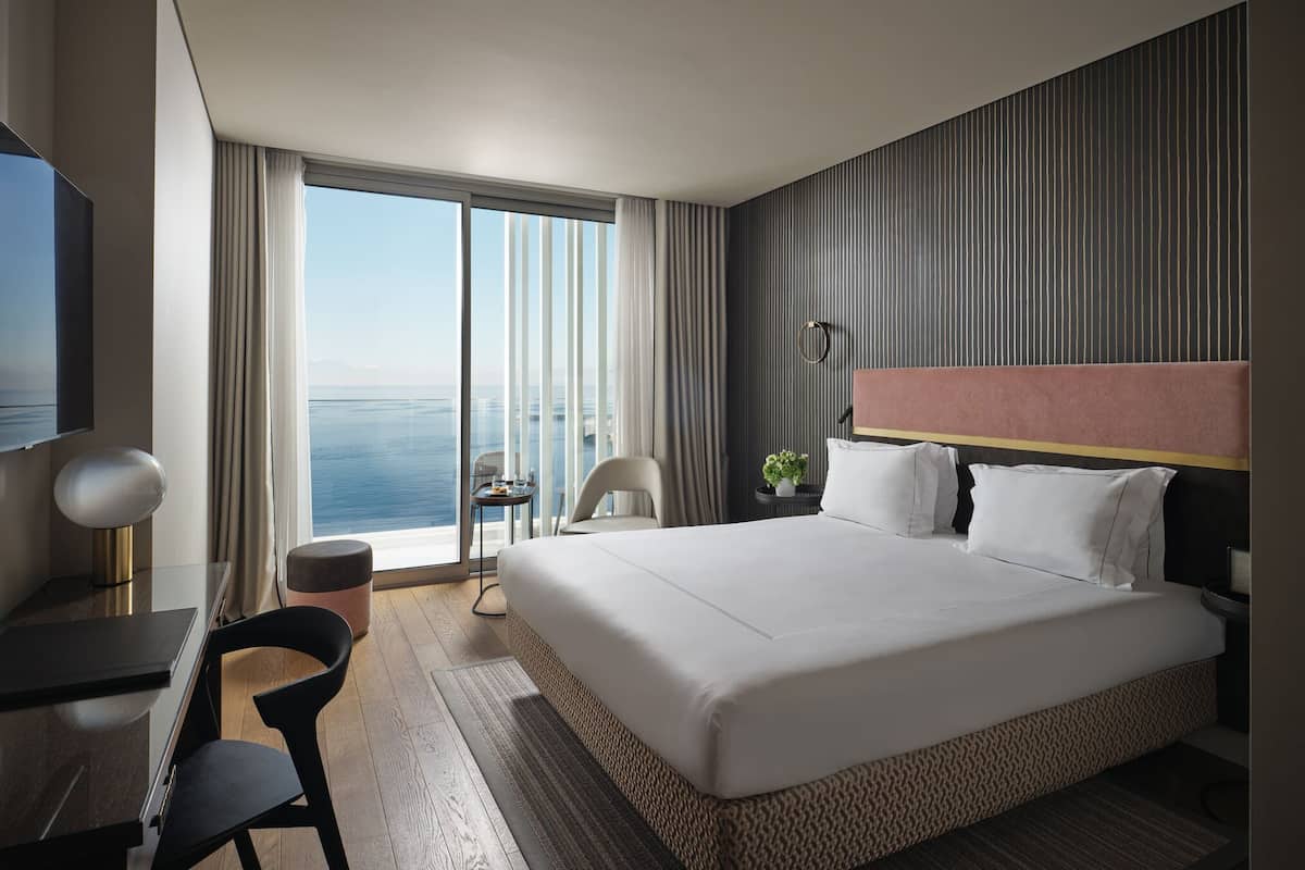 A hotel room with a large bed, a desk, a chair, a TV, and a view of the ocean.