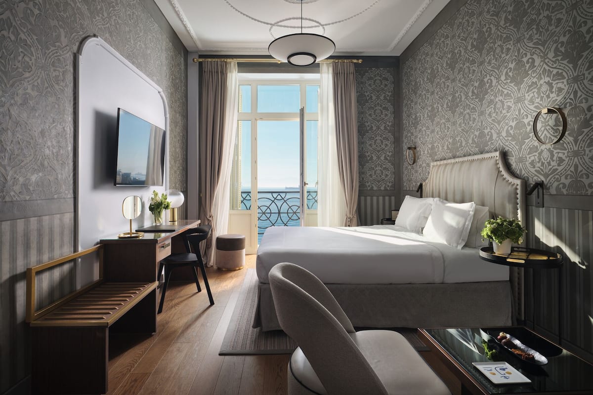 A hotel room with a bed, a desk, a chair, a TV, and a view of the sea.