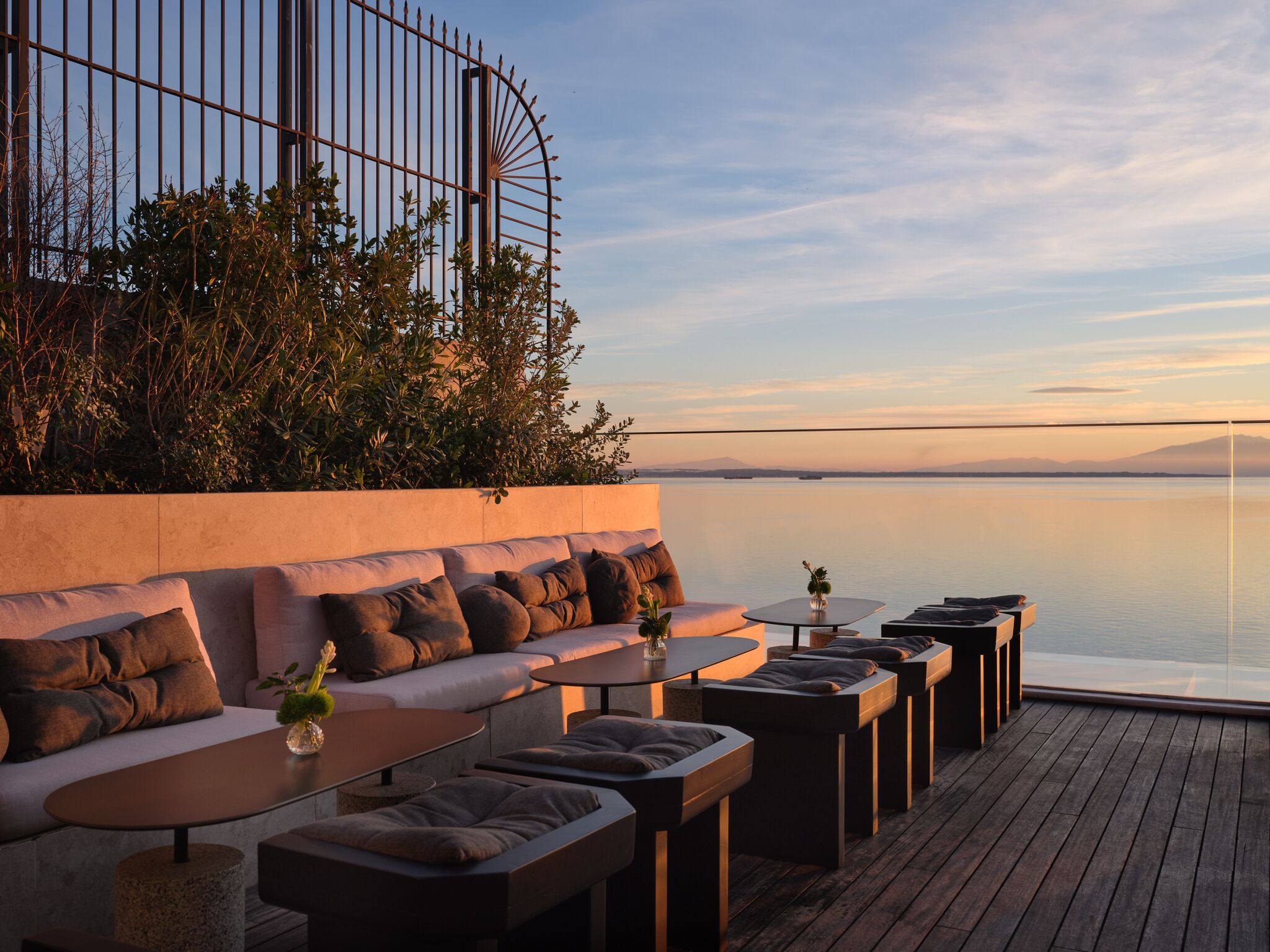 A deck with a sofa, ottomans, and tables, overlooking a calm body of water and distant mountains.