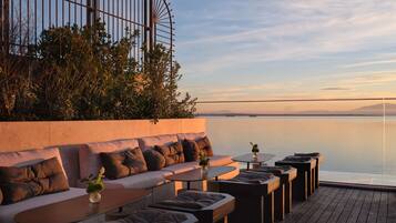 A deck with a sofa, ottomans, and tables, overlooking a calm body of water and distant mountains.