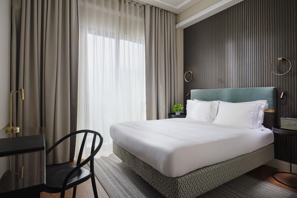 A hotel room with a bed, a desk, a chair, a small plant, and a wall with vertical lines.