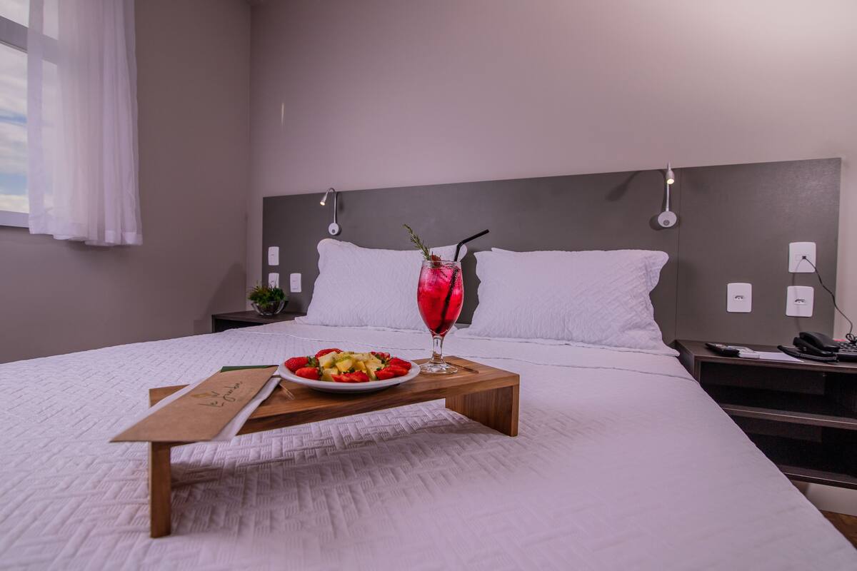 A neatly made bed with white linens, a bedside table with a phone and a lamp, and a tray with a fruit salad and a drink.