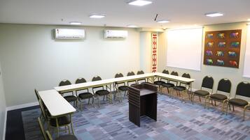 Un salón de conferencias con una larga mesa, sillas, un atril, unidades de aire acondicionado, una pantalla de proyector y una decoración de animales de colores en la pared.