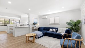 Un salon moderne avec un canapé bleu foncé, une table basse en bois et un fauteuil bleu. On y trouve également un coin cuisine avec des meubles blancs et une fenêtre équipée de stores.