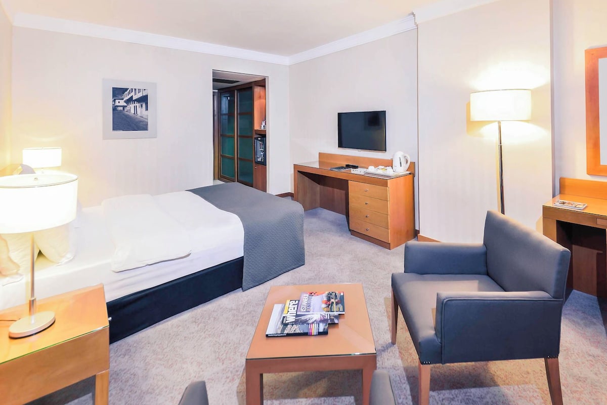 A hotel room with a bed, desk, two chairs, a TV, and a lamp.