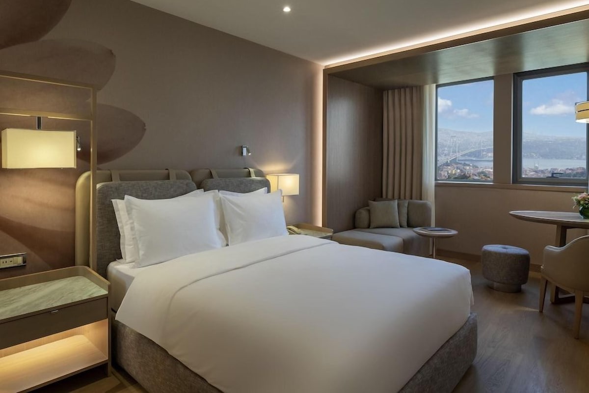 A modern hotel room with a large bed, a desk, a chair, and a view of a cityscape.