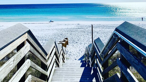 Une promenade en bois qui mène à une plage où l’eau est d’un bleu limpide et le ciel est radieux.