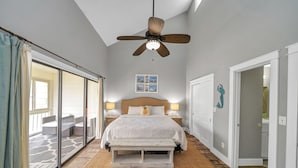 Une chambre dotée d’un grand lit, de deux tables de chevet avec des lampes, d’un ventilateur de plafond et offrant une vue sur un espace extérieur grâce à une porte-fenêtre coulissante.