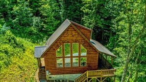 A wooden cabin with a large window, a deck, and a staircase leading to a second level.