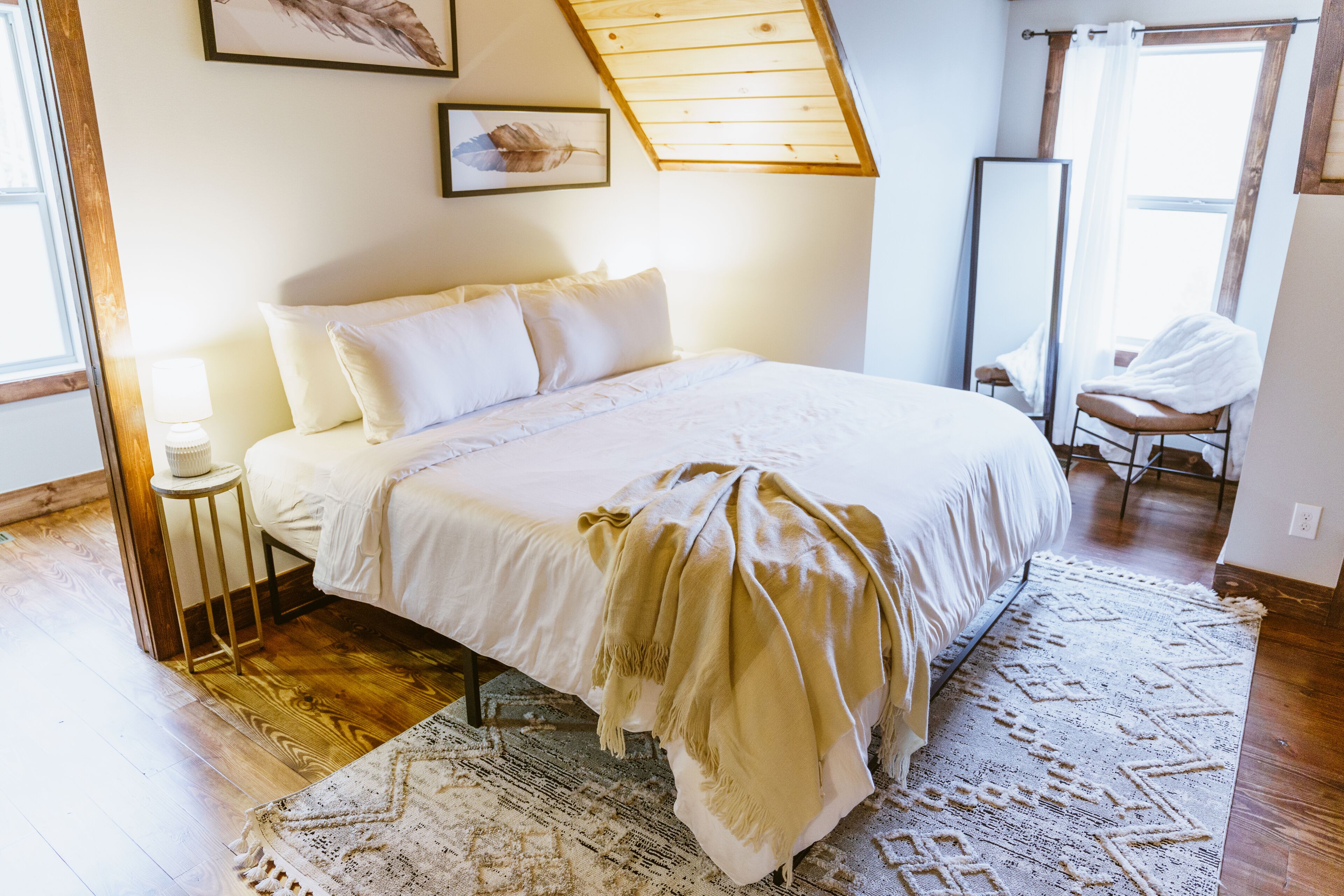 Ein Schlafzimmer mit einem großen Bett, Holzboden, einem stehenden Spiegel und einem Fenster mit Vorhängen.