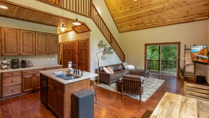 A spacious living area with a wooden staircase, a kitchen with wooden cabinets, a dining table, and a large window offering a view of the outdoors.