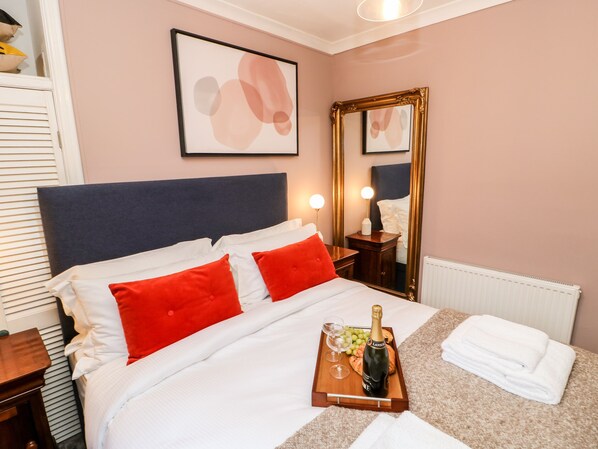 A neatly made bed with red and white pillows, a bedside table with a bottle of champagne and glasses, and a mirror on the wall. - Jaipur House (Richmond)