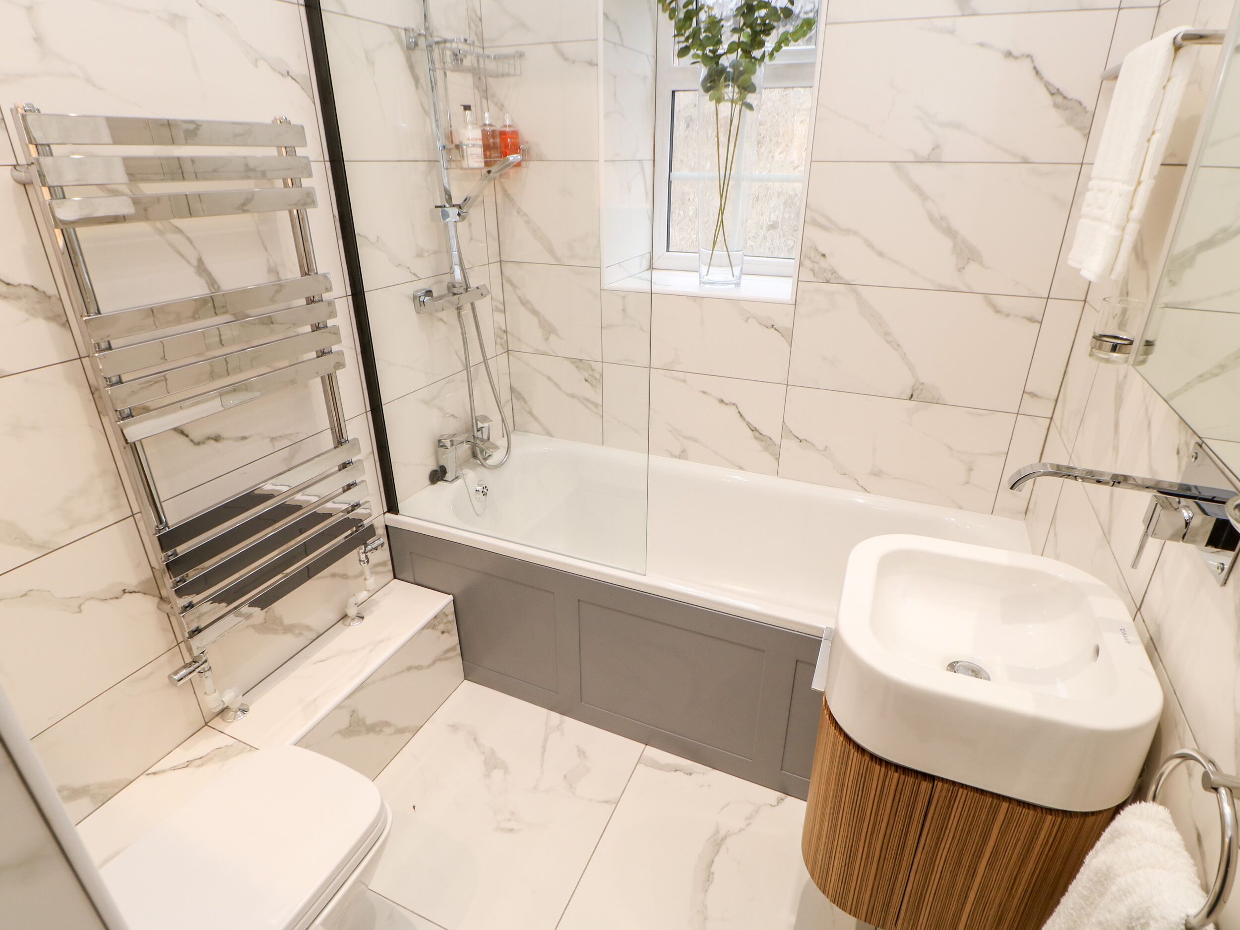 Ein modernes Badezimmer mit einer Badewanne, einer Toilette und einem Waschbecken. Ein Duschbereich mit einer Glasabtrennung und ein Fenster über der Badewanne.