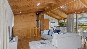 Un salon chaleureux avec un plafond en bois, un canapé gris agrémenté d’un magazine et une vue sur l’extérieur grâce à une grande fenêtre.