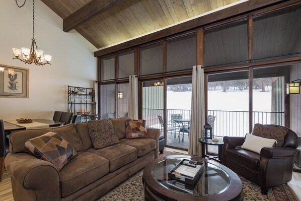 A spacious living room with a brown leather sofa, a glass coffee table, and a fireplace. There is a dining area with a table and chairs, and a large window offering a view of the outdoors.