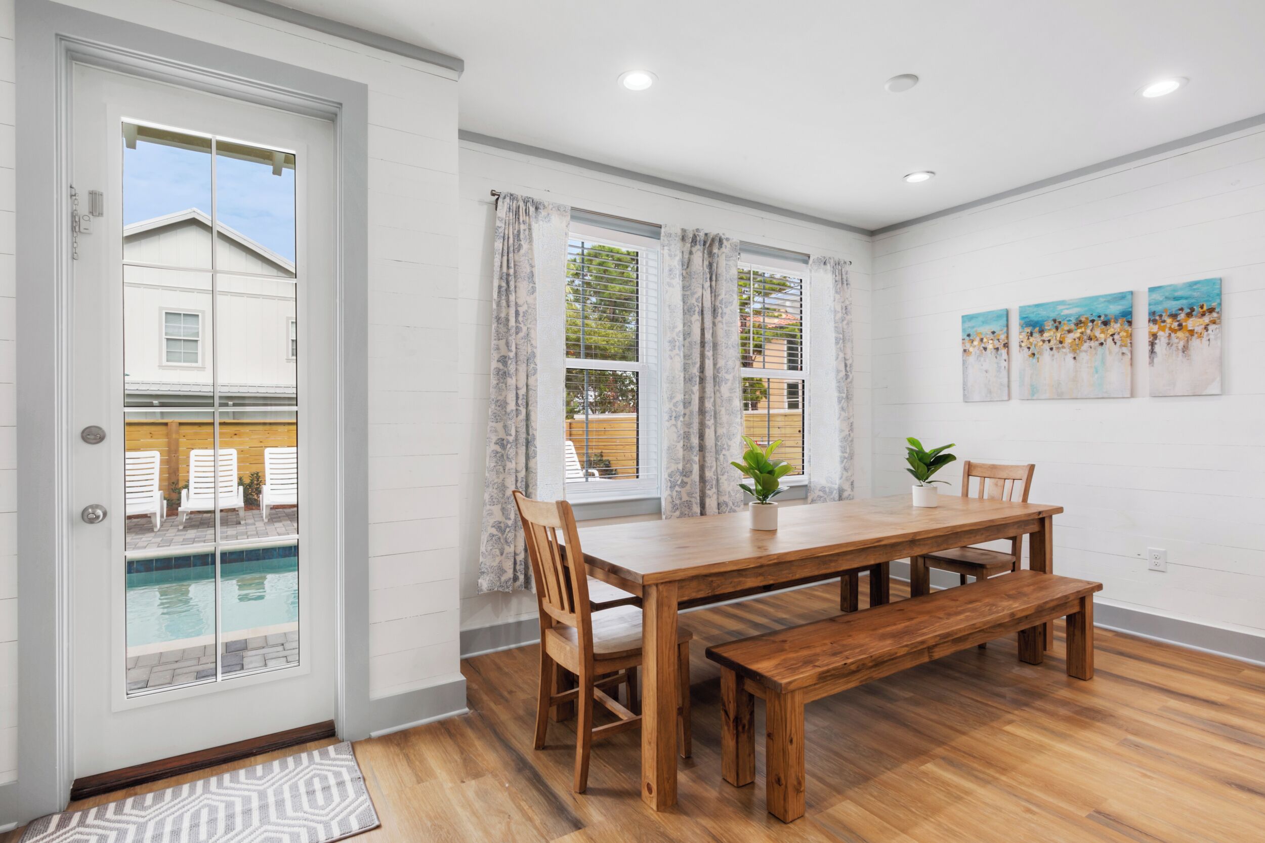 A dining area with a wooden table, bench, and chairs, a potted plant, and a view of a swimming pool through a glass door.
