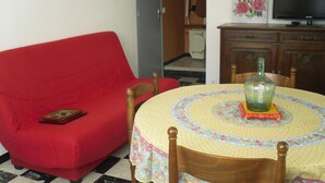 A red sofa, a wooden dining table with a floral tablecloth, a bottle, and a television on a wooden cabinet. - Village house near spain, sea, lake, shops nearby (Le Boulou)