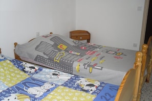 Un lit simple avec une tête de lit représentant un paysage urbain et une couette aux motifs de personnages de dessins animés, une table de chevet en bois et une porte dans une pièce aux murs blancs.