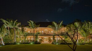 A large, well-lit building with a symmetrical facade, surrounded by palm trees and a well-maintained lawn. - One-of-a-kind Villa With Open Spaces and Amazing Views in Luxury Beach Resort (Punta Cana)