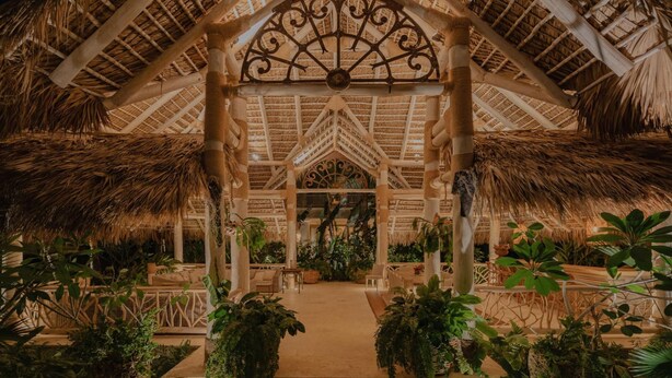 Une construction au toit de chaume, dotée d’un élément décoratif central, entourée de verdure et d’espaces aménagés pour s’asseoir.