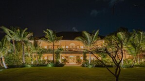 Un grand bâtiment bien éclairé, doté d’une façade symétrique, entouré d’une végétation luxuriante et de palmiers.