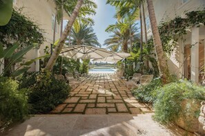 A paved pathway leading to a pool area with wicker chairs and umbrellas, surrounded by lush tropical plants and palm trees.