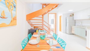 A dining area with a wooden table set for six, a staircase, and a kitchen with white cabinets. - IT28 Triplex 4 Suítes 100m da Praia (Camaçari)