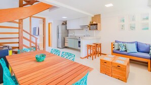 A modern kitchen with white cabinets, a microwave, and a refrigerator. There is a wooden dining table with chairs and a blue sofa in the living area. - IT28 Triplex 4 Suítes 100m da Praia (Camaçari)
