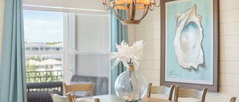 A dining table with a glass vase and white flowers, wooden chairs, a chandelier, and a framed seashore painting.