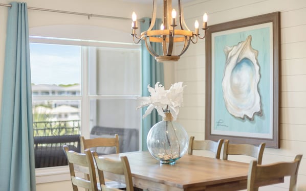 A dining table with a glass vase and white flowers, wooden chairs, a chandelier, and a framed seashore painting.