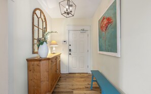 A hallway with a wooden cabinet, a white vase with flowers, a wall-mounted lamp, a door with a security lock, and a blue bench. - New Rental in Seacrest Beach Resort, stunning views of Lagoon Pool, 3 bikes! (Panama City Beach)