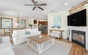 A spacious living room with a white sofa, a wooden coffee table, a fireplace, and a television mounted on the wall.