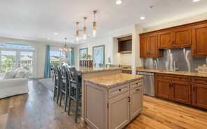 A modern kitchen with wooden cabinets, a stainless steel refrigerator, and a dining area with a white sofa and chairs. - New Rental in Seacrest Beach Resort, stunning views of Lagoon Pool, 3 bikes! (Panama City Beach)