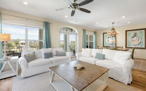 A spacious living room with white sofas, a wooden coffee table, and a view of the outdoors through large windows. - New Rental in Seacrest Beach Resort, stunning views of Lagoon Pool, 3 bikes! (Panama City Beach)
