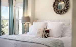 A bedroom with a bed, pillows, a lamp, a mirror, and a window with blinds.