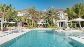A pool area with lounge chairs and umbrellas, surrounded by palm trees and a building with a red roof. - Luxurious Fully-staffed Villa With Amazing View in Exclusive Golf Beach Resort (Punta Cana)