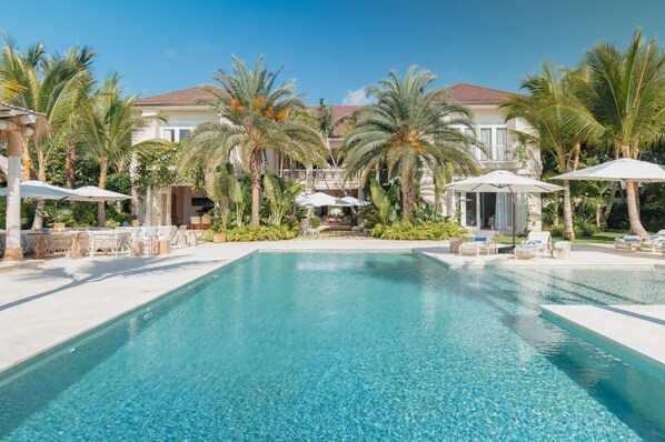 A pool area with lounge chairs and umbrellas, surrounded by palm trees and a building with a red roof.