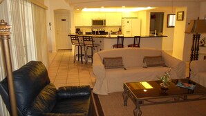 A living room with a black leather sofa, a glass coffee table, and a white sofa with patterned cushions. A kitchen is visible in the background with a bar and chairs.