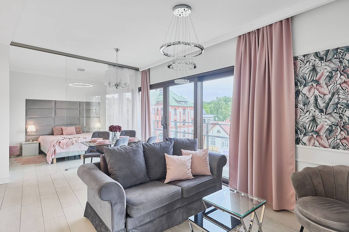 Una sala de estar moderna con un sofá gris, una mesa de centro de vidrio y un ventanal con cortinas.