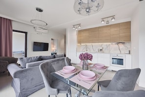 A modern living room with a glass dining table, grey sofas, and a kitchen with wooden cabinets and a marble countertop. - Apartamenty Swinoujscie - Kormoran (Swinoujscie)