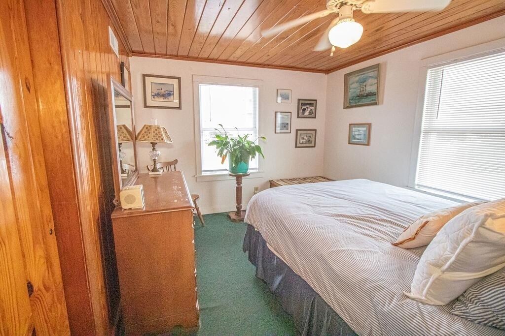 Un dormitorio con techo de madera, una cama con colcha a rayas, un aparador, una lámpara y un ventanal con persianas.